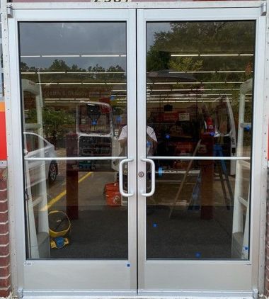 Double glass doors with silver frames, reflecting a store interior and exterior. 