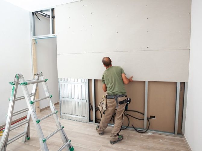 Worker installing drywall in a room with a ladder and exposed wall framing