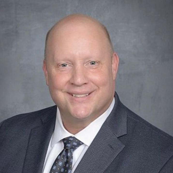 Bald man in a suit and tie smiles at the camera against a gray backdrop.