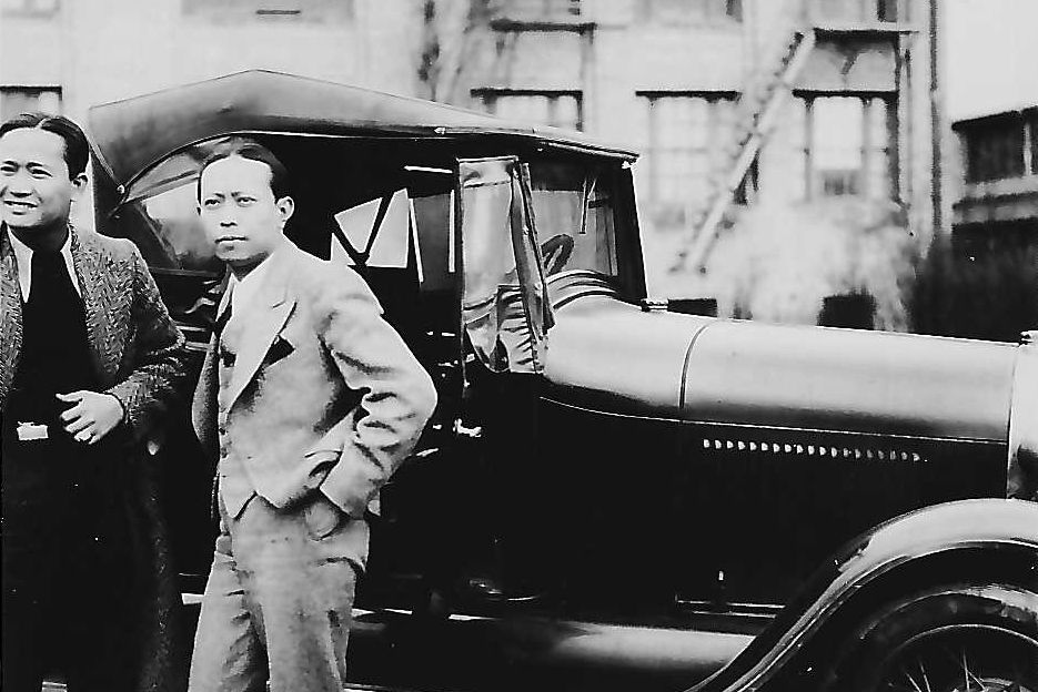 A black and white photo of two men standing next to a car