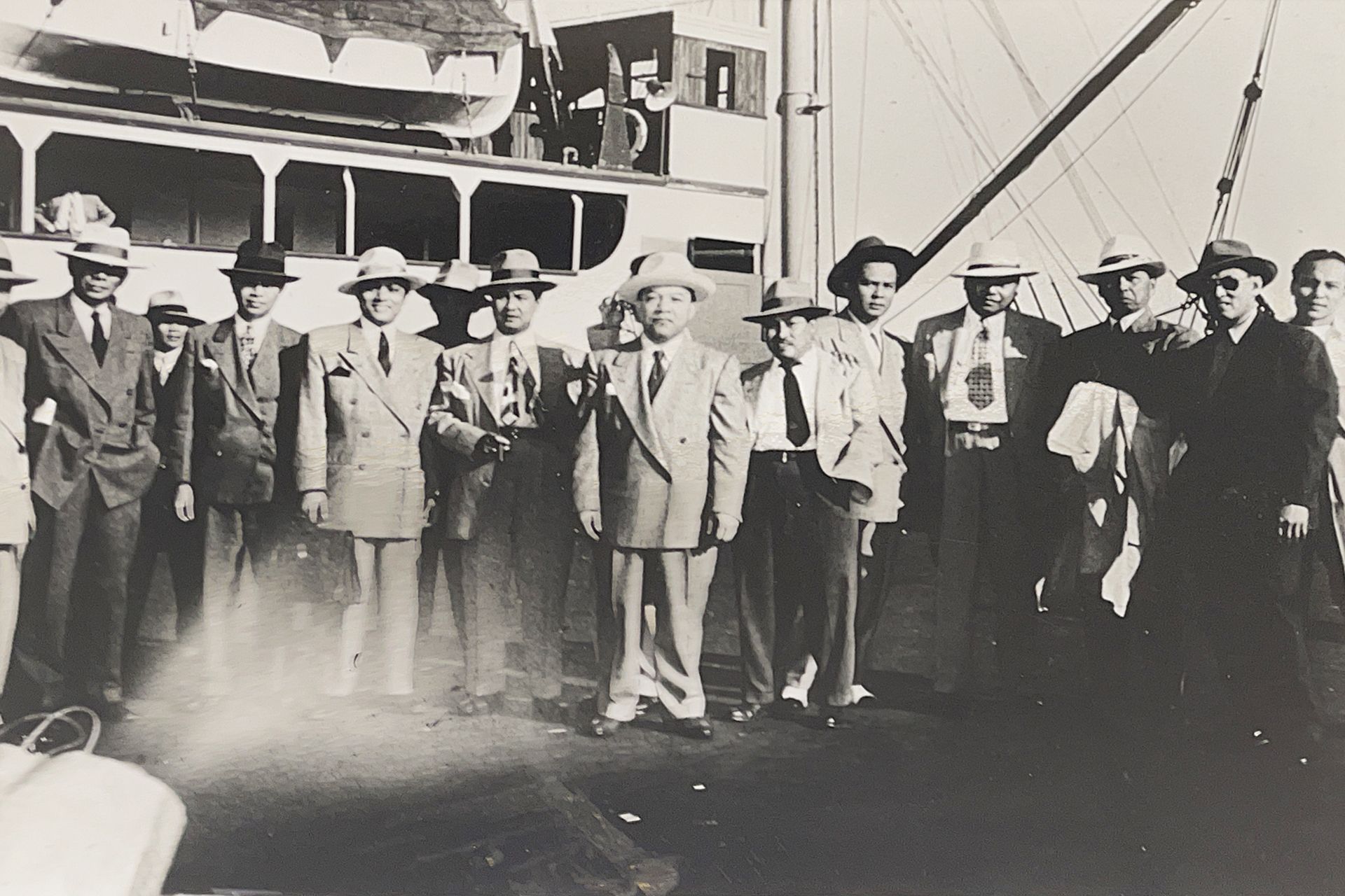 A black and white photo of a group of men in suits and hats in front of a boat