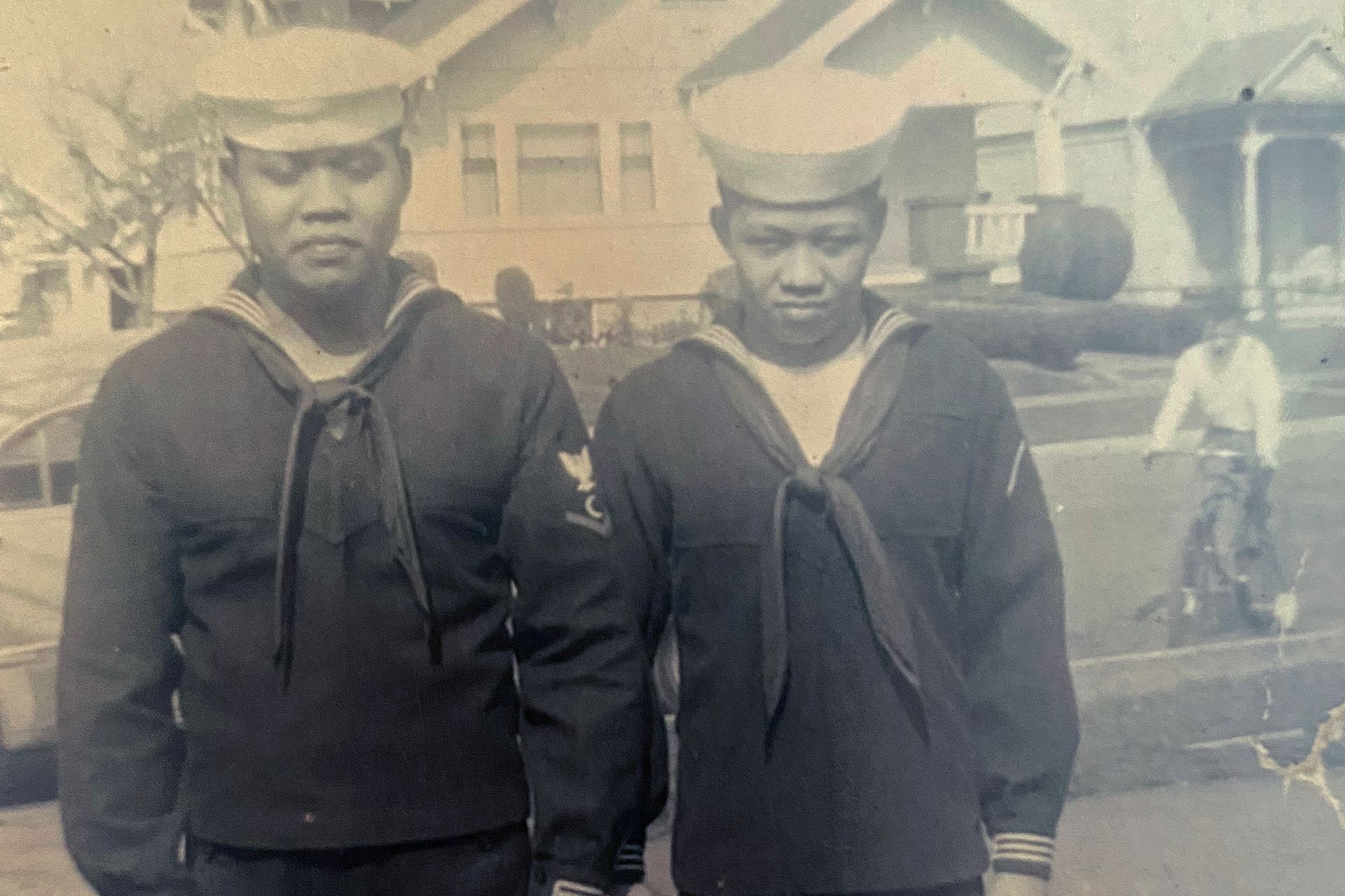 Two young men in sailor uniforms stand next to each other in front of a house