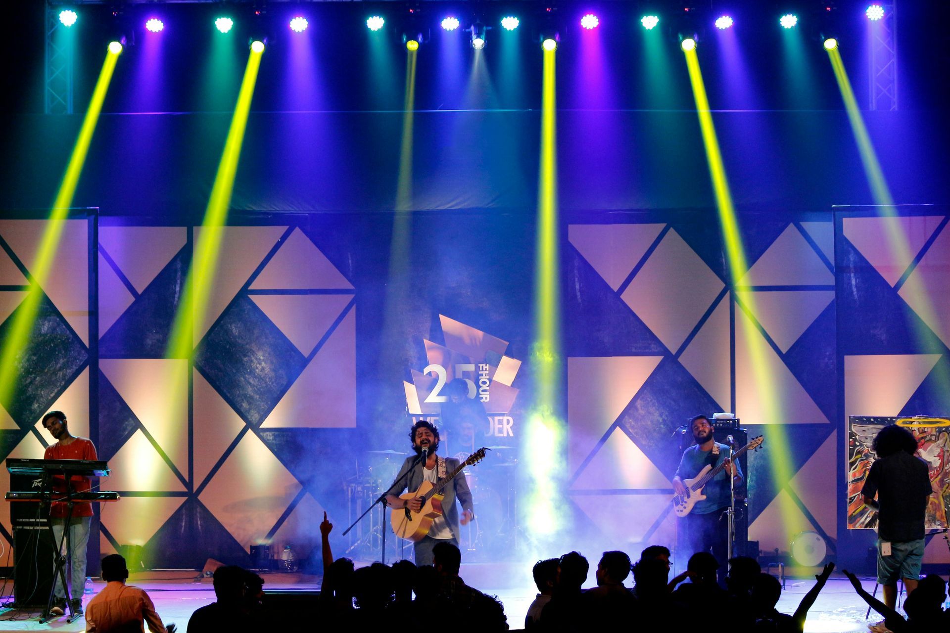 Live band performing on a colorful stage with bright blue and yellow lights and geometric backdrop