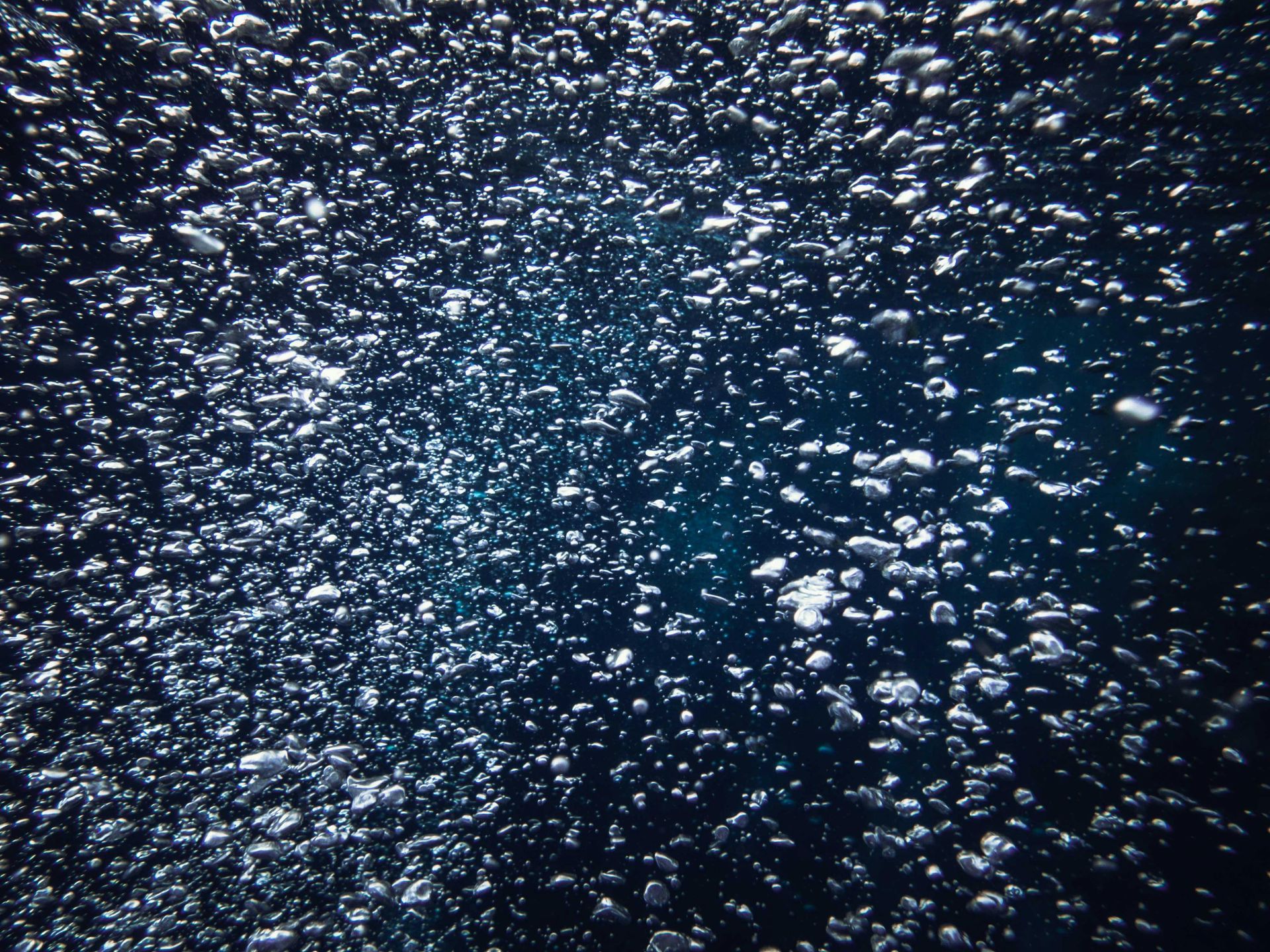 A dense cluster of bubbles rising through deep blue, dark water.