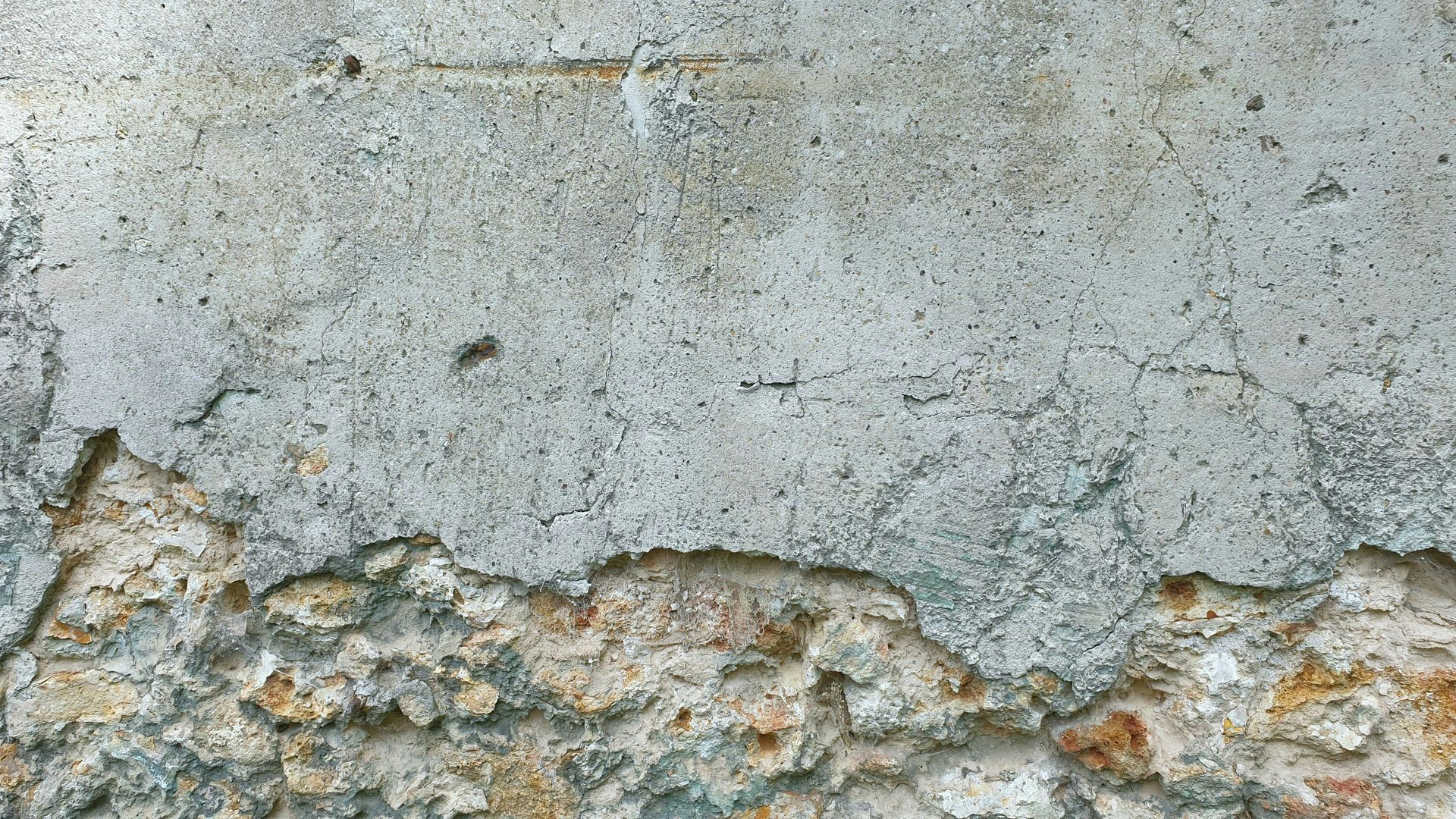 A weathered wall with peeling gray plaster revealing rough, tan-colored brick underneath.