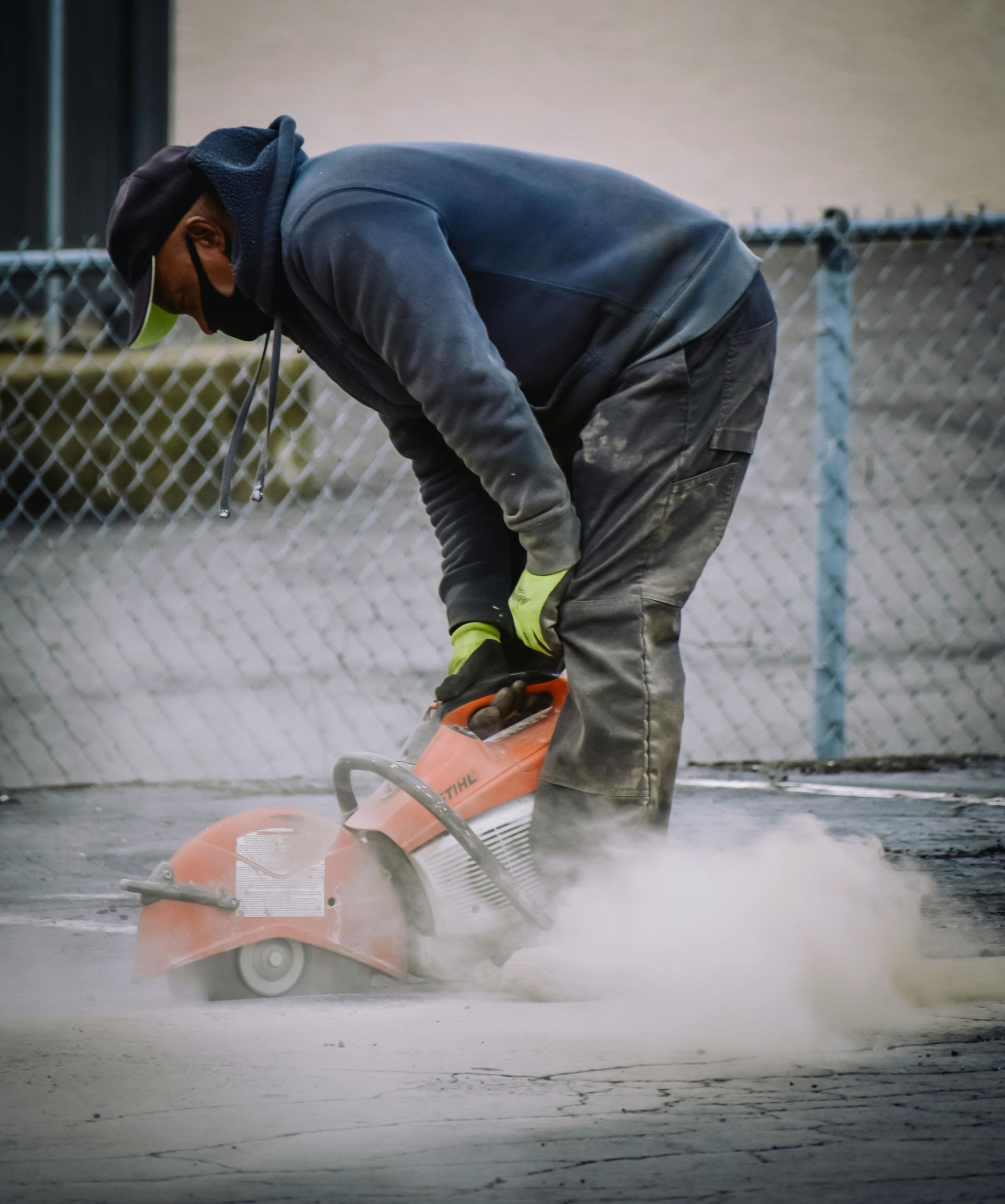 Worker using a cut-off saw on pavement, with dust and debris around the blade.