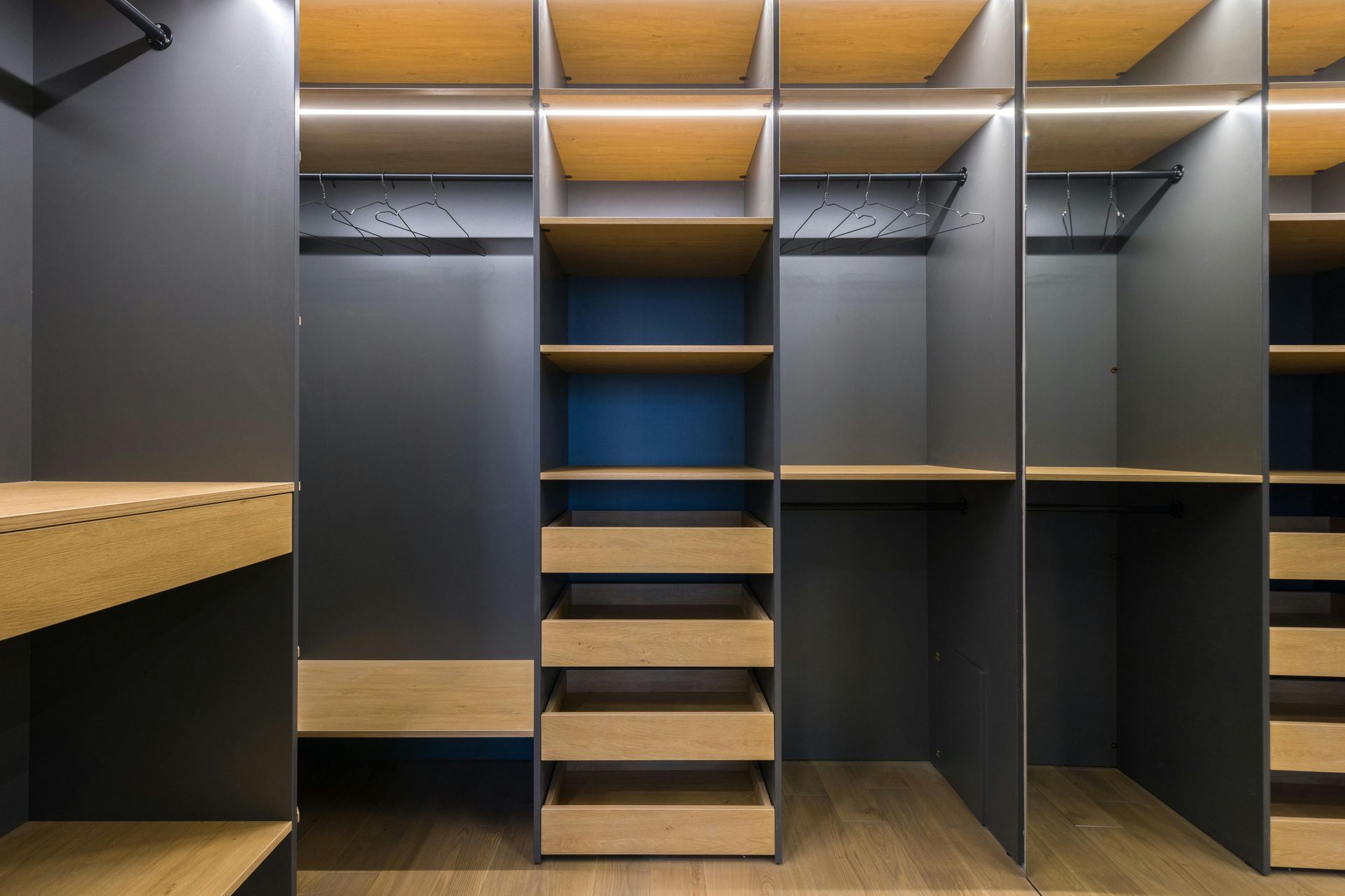 Empty walk-in closet with dark gray walls, wood shelves, drawers, and hanging rods; illuminated by overhead lights.