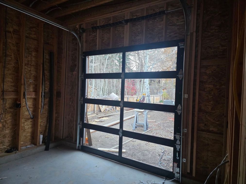 Partially open black garage door in an unfinished wood-framed interior, showing a snowy yard outside