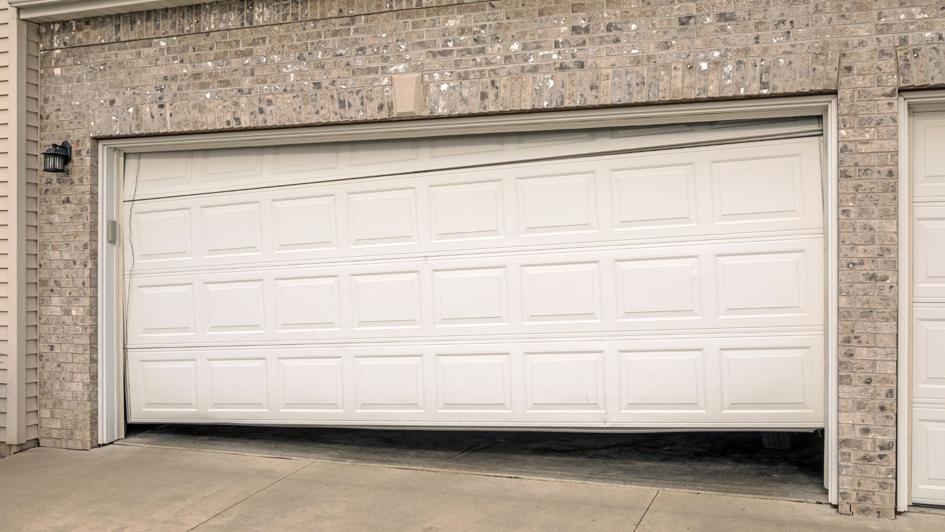 A white, multi-panel garage door is crooked, significantly raised on the left side, and hanging needing emergency garage door repair