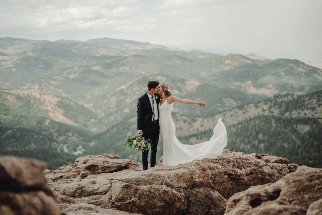 A bride and groom are kissing on top of a mountain.