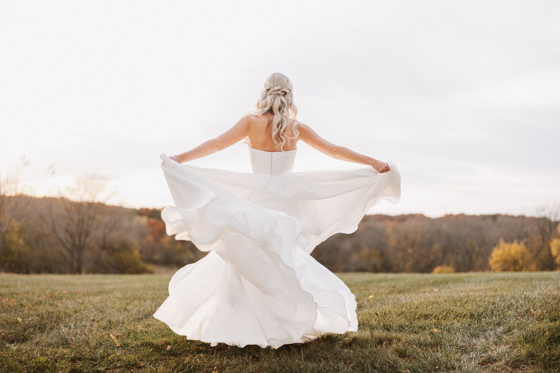 A bride in a white dress is standing in a field with her arms outstretched.