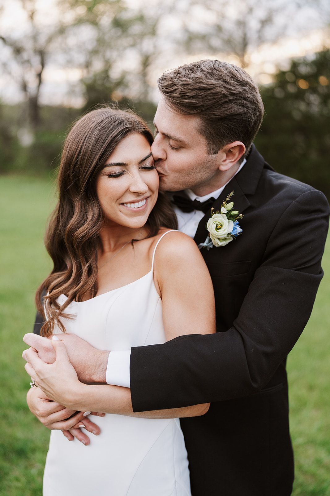 A bride and groom are posing for a picture and the groom is kissing the bride on the cheek.