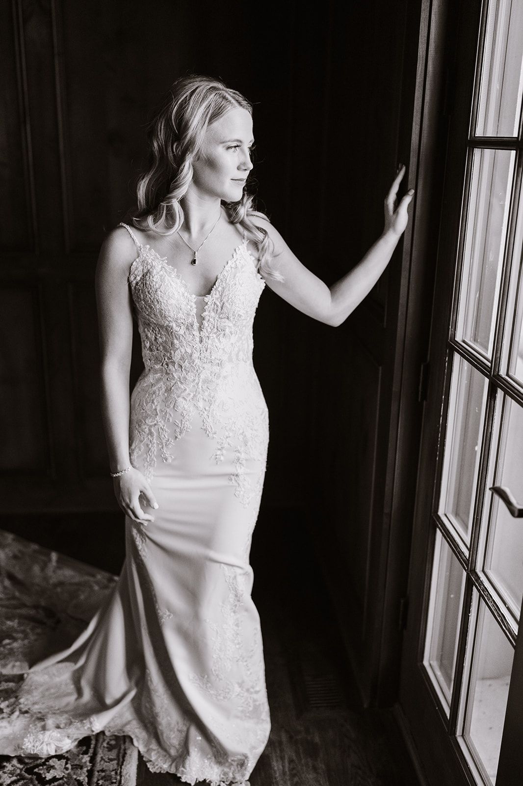 A black and white photo of a bride in a wedding dress standing in front of a window.