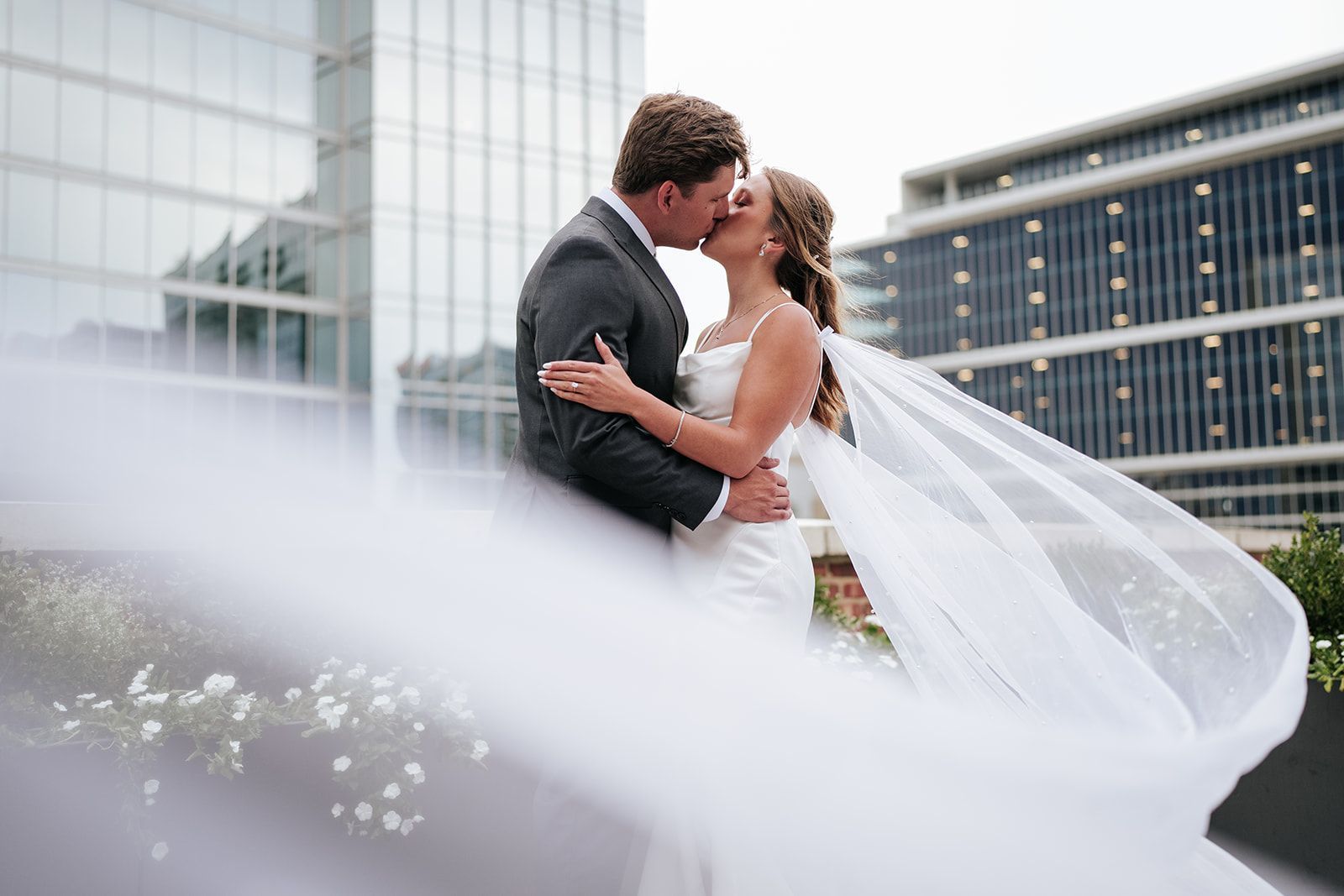 A bride and groom kissing in front of a building with a veil blowing in the wind.
