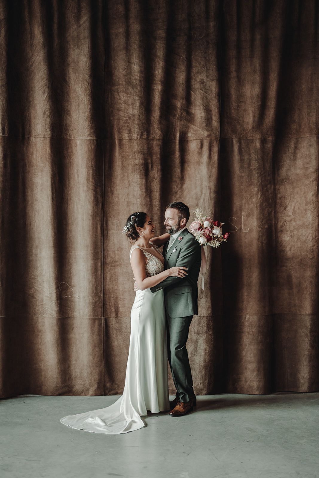 A bride and groom are standing next to each other in front of a brown curtain.