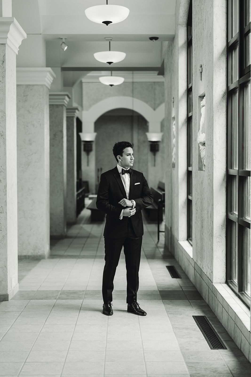 A man in a tuxedo is standing in a hallway next to a window.