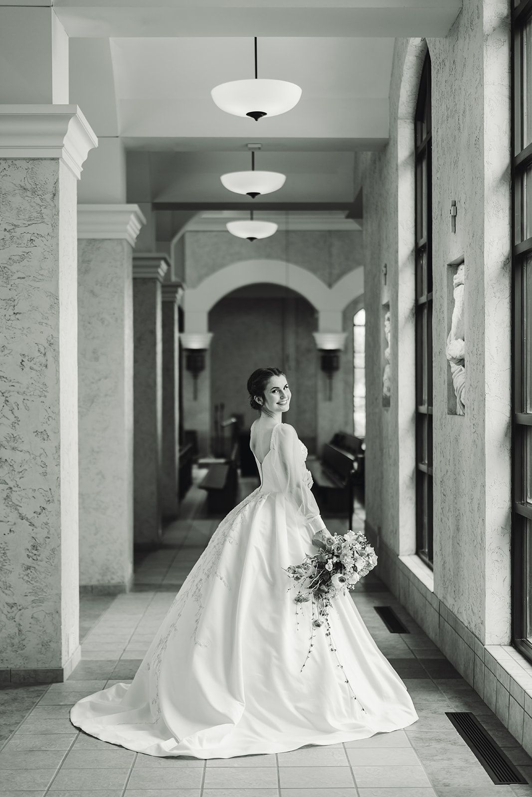 A bride in a wedding dress is standing in a hallway holding a bouquet of flowers.