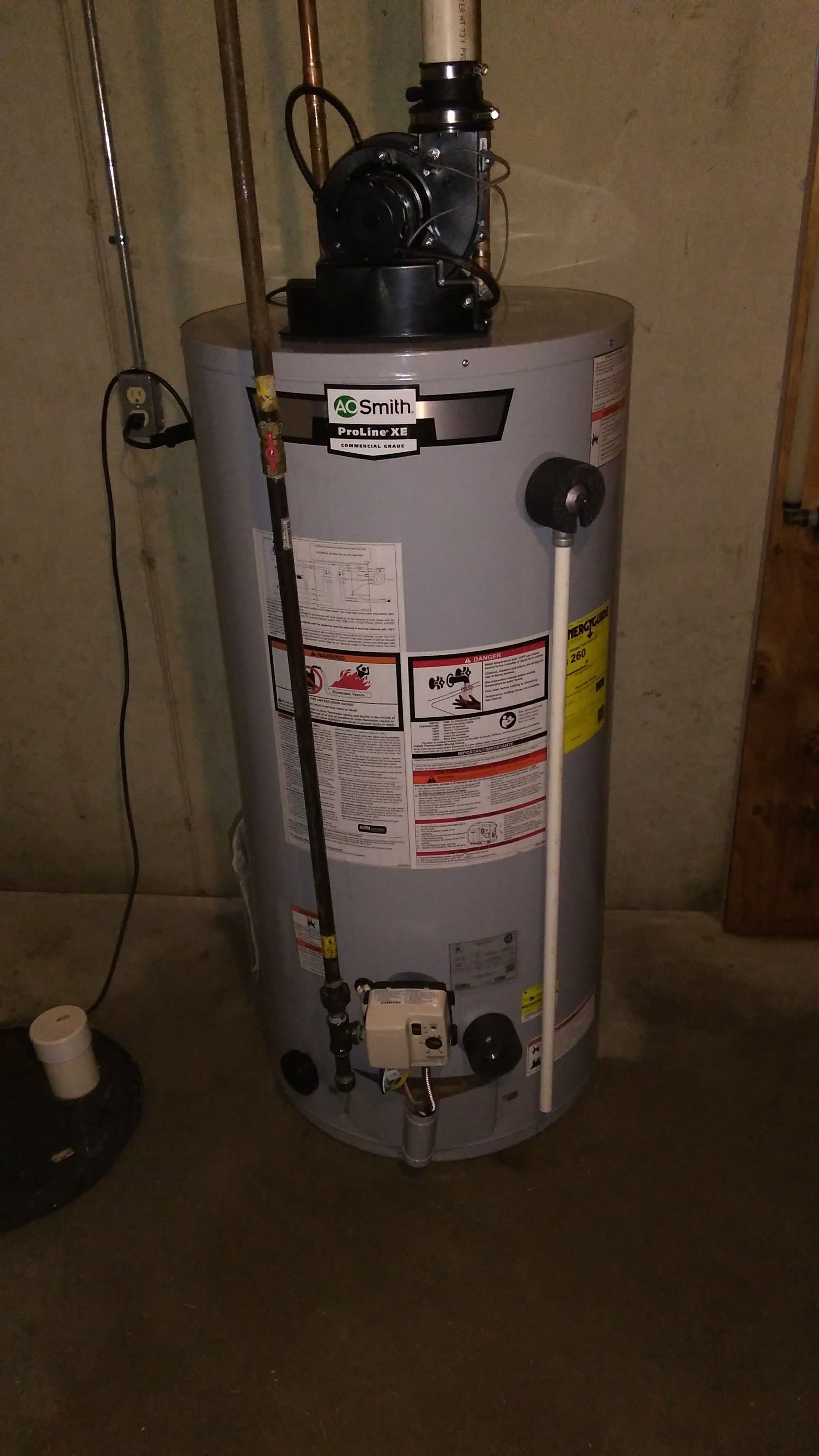 A grey, cylindrical gas water heater stands on a concrete floor in a basement, featuring a power vent blower on top.
