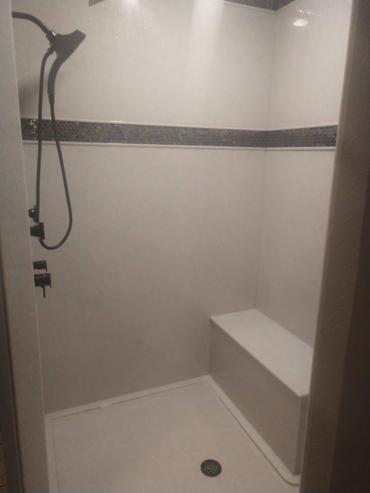 A white shower stall featuring a handheld showerhead, a built-in bench, and a dark horizontal decorative tile border.