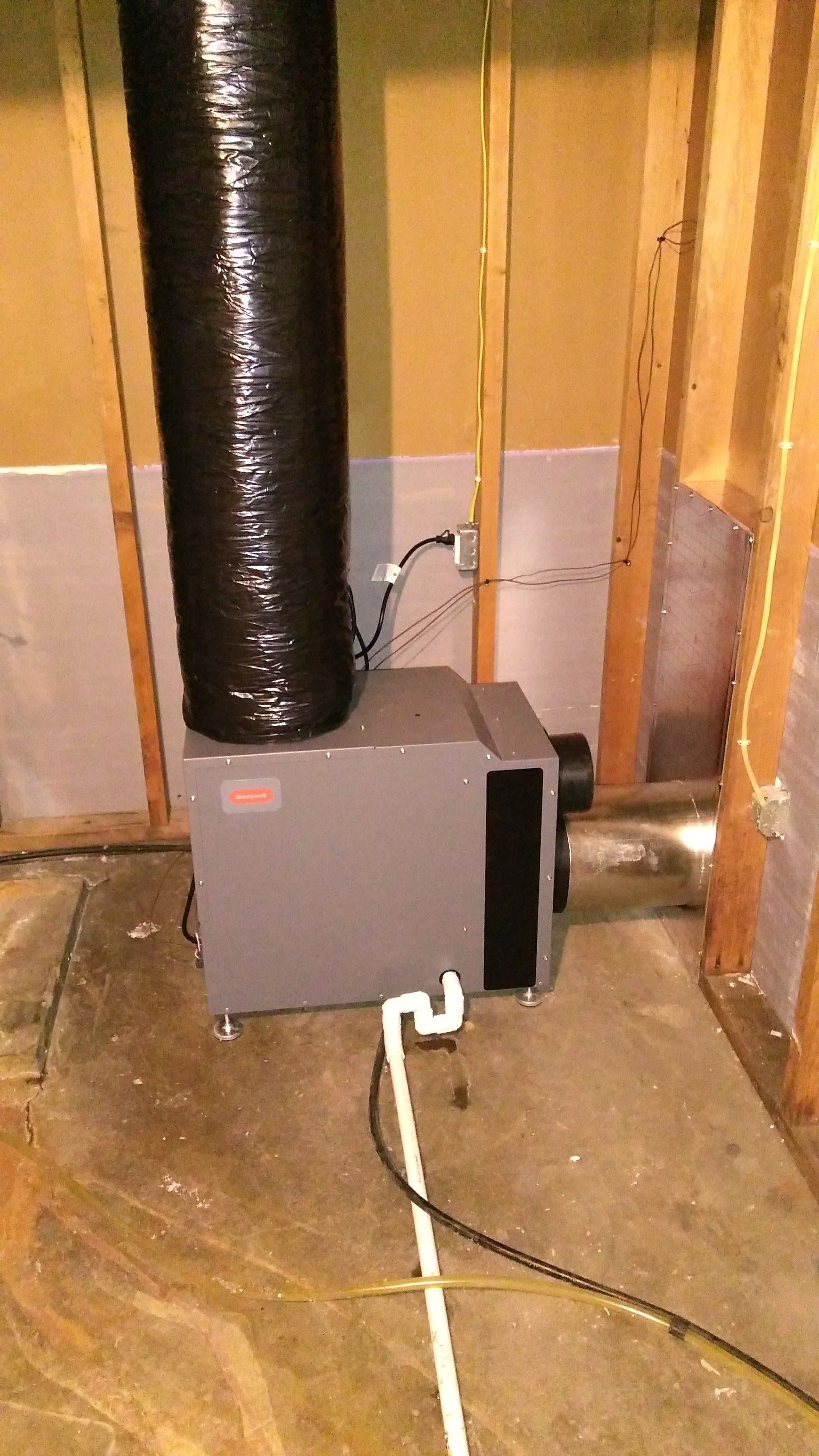 A gray dehumidifier with a black flexible duct sits on a concrete floor between unfinished wooden wall studs.