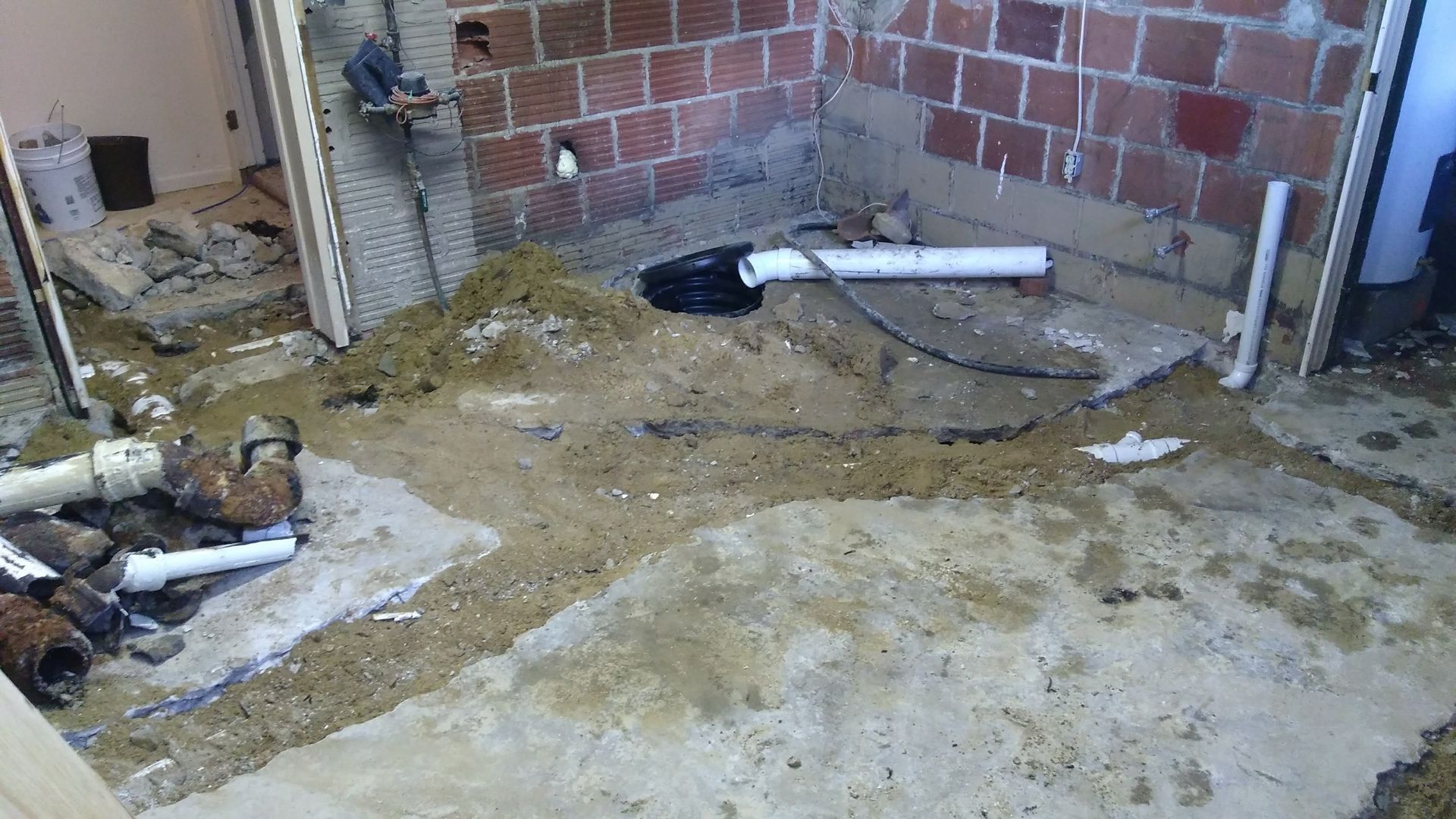 A construction site showing exposed brick walls, dirt flooring, and PVC plumbing pipes during a home renovation.