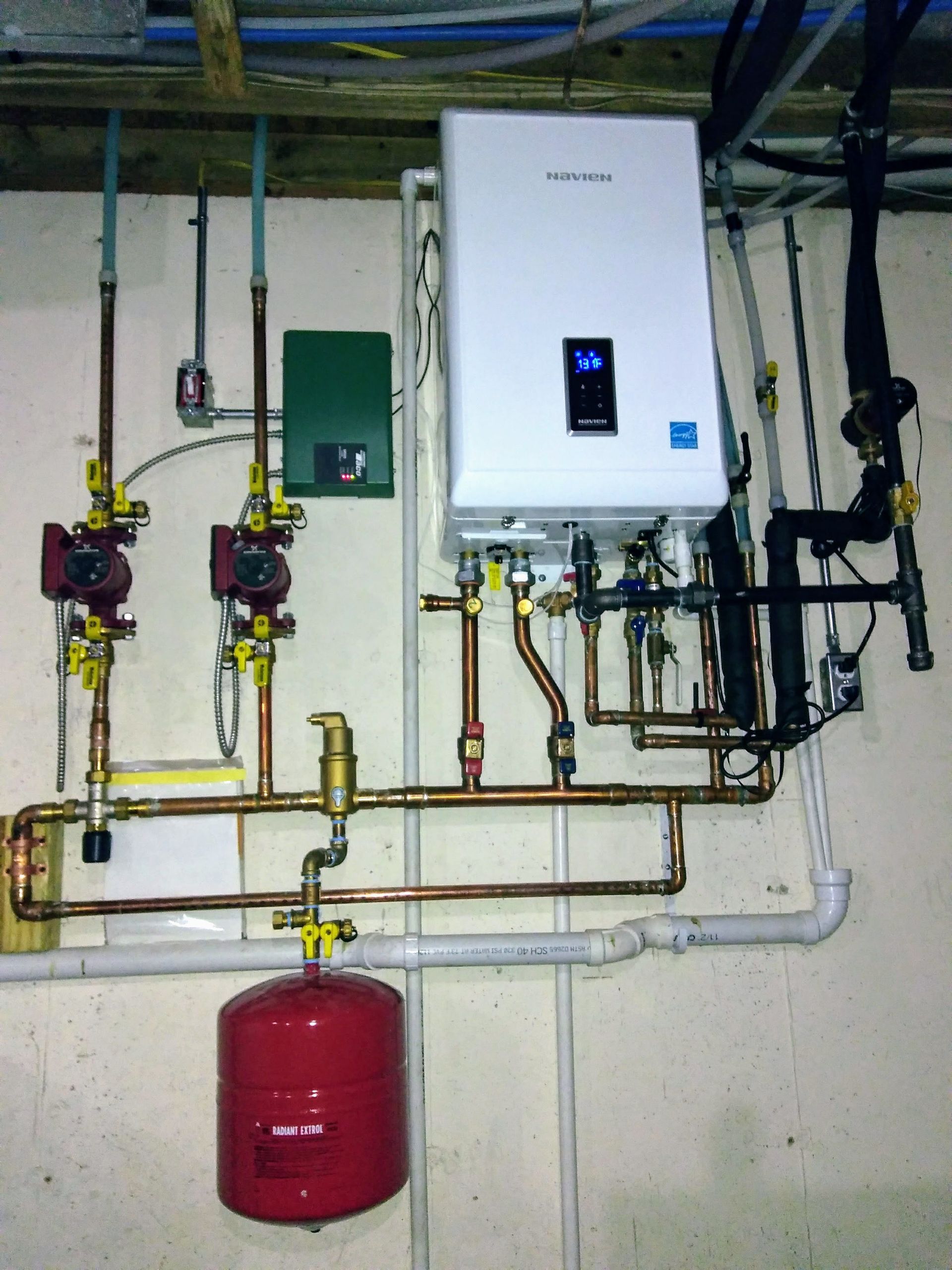 Wall-mounted Navien tankless water heater with copper piping, circulator pumps, and a red expansion tank on a basement wall.