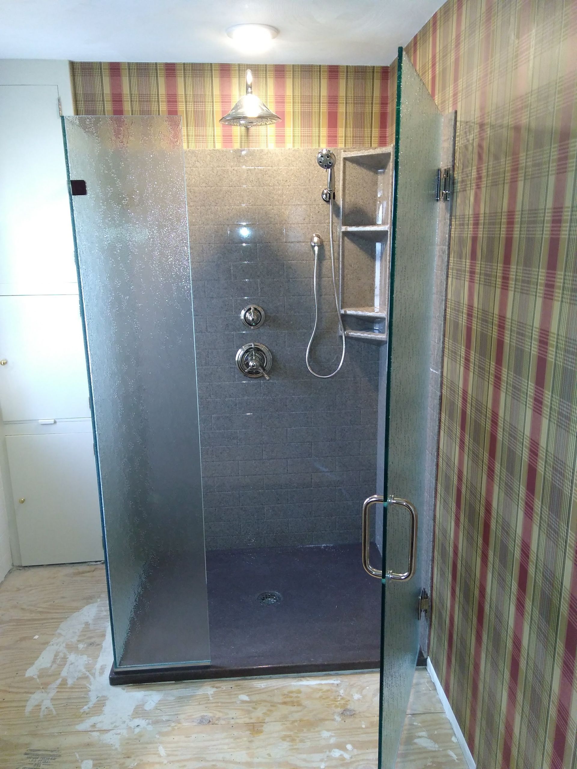 A glass-enclosed shower with gray tiled walls, a handheld showerhead, built-in shelving, and striped wallpaper exterior.