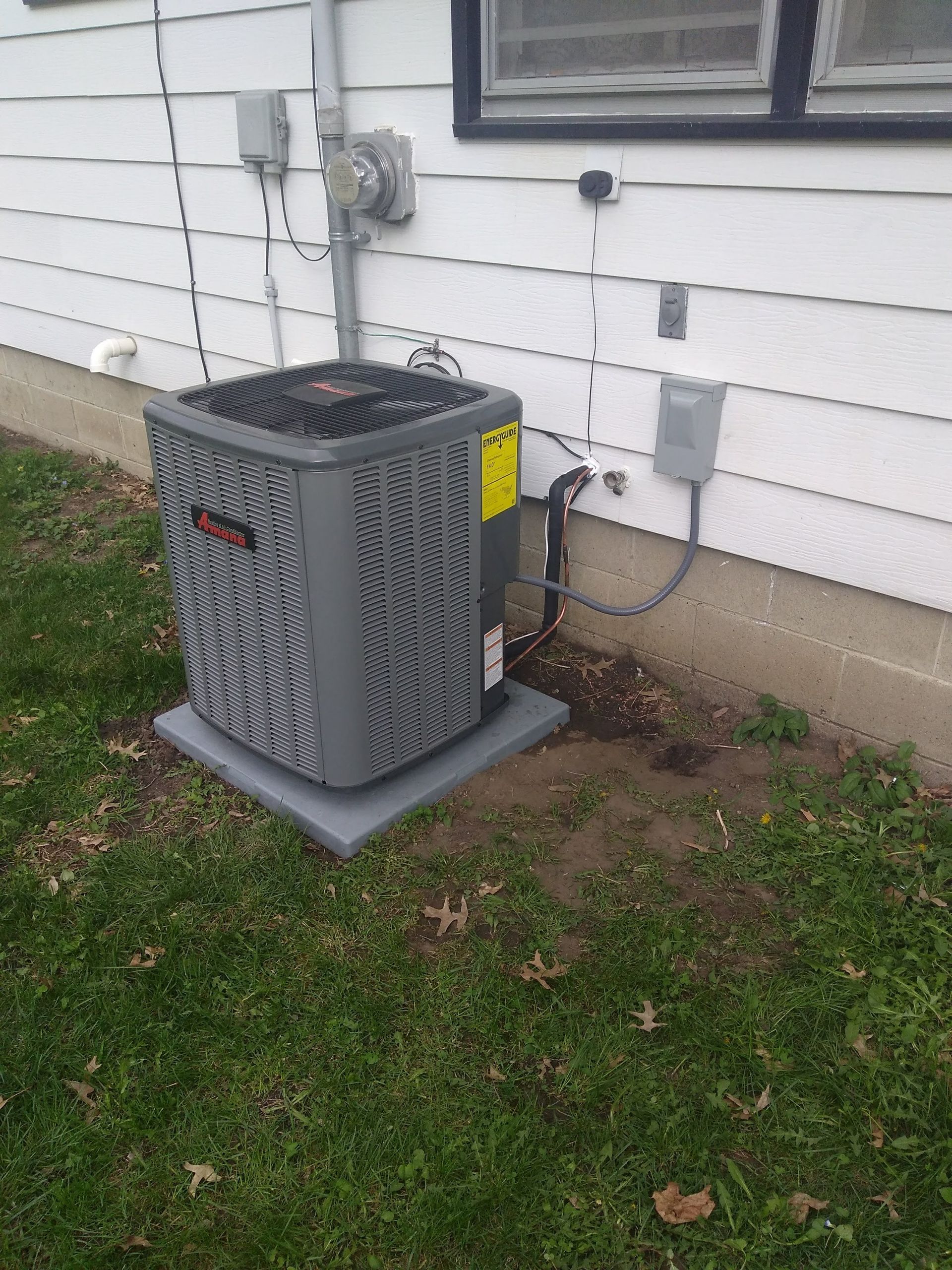 A grey outdoor air conditioning unit sits on a concrete pad against the white siding of a house on a grassy lawn.