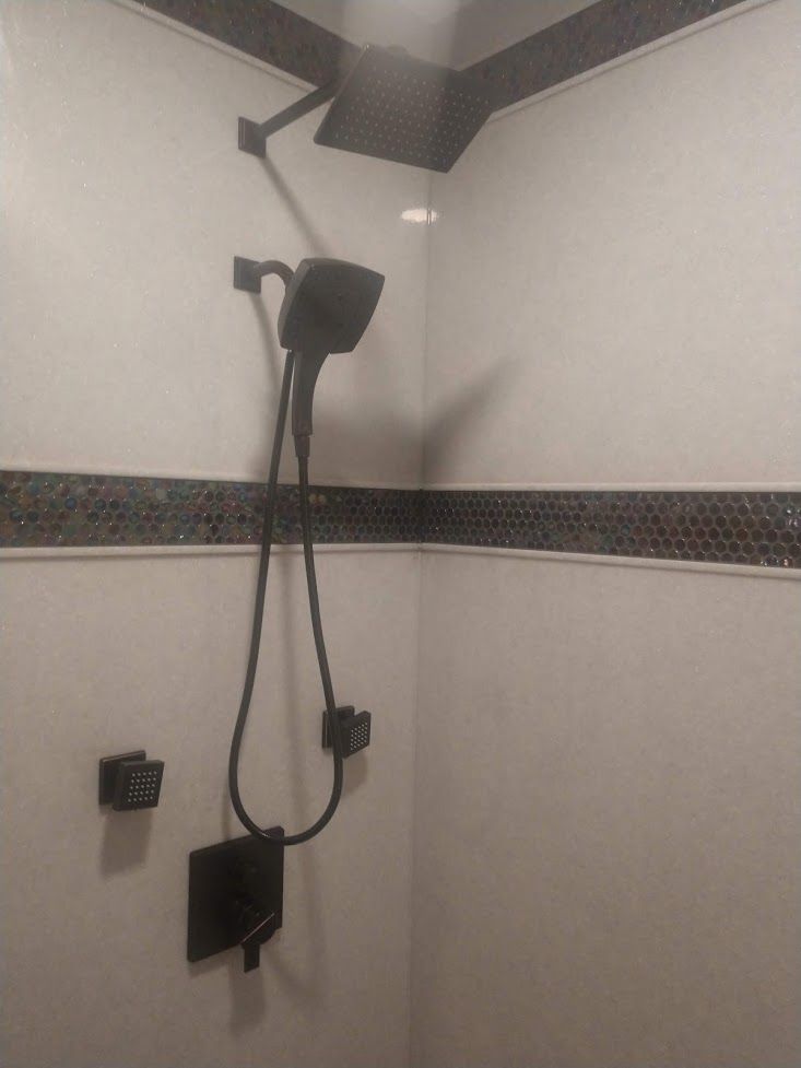 A modern shower with a matte black rainfall head, a handheld sprayer, and a control handle against white, tiled walls.
