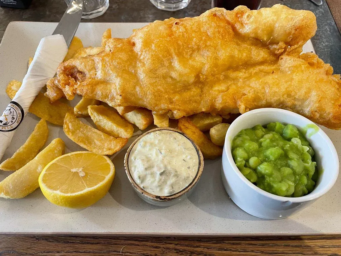 A plate of fish and chips with peas and sauces on a table.