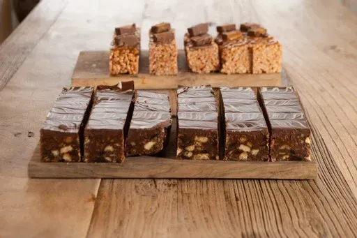 A wooden cutting board topped with a variety of brownies on a wooden table.