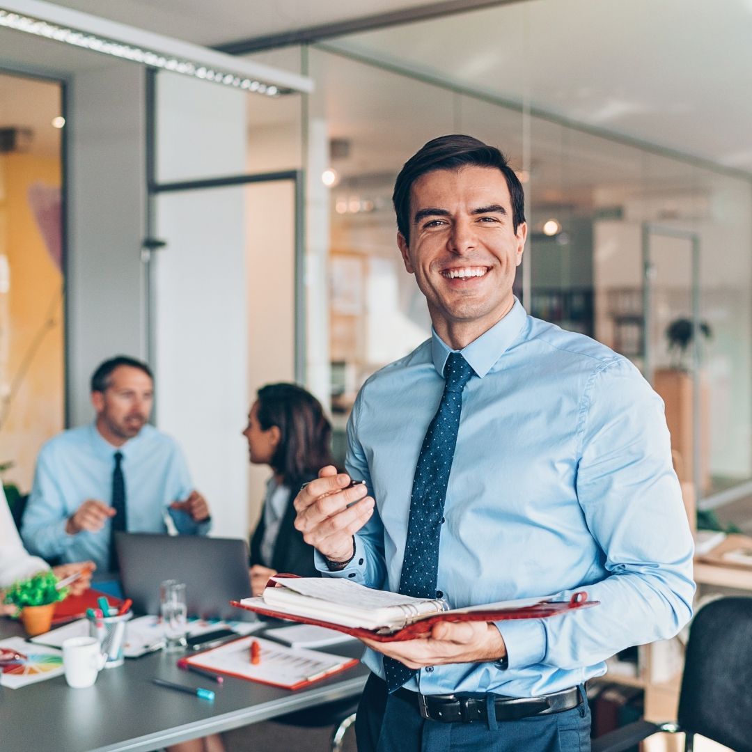 Gestão Administrativa Síndico Profissional Empresário sorridente segurando um caderno no escritório com equipe trabalhando ao fundo.