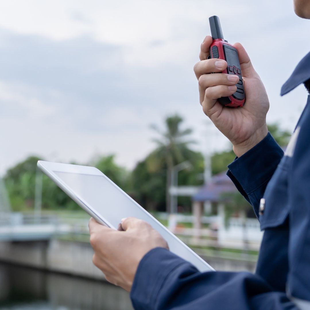 Gestão Manutenção Predial Pessoa de uniforme azul segurando um tablet e um walkie-talkie ao ar livre, perto de uma estrutura - manutenção predial.