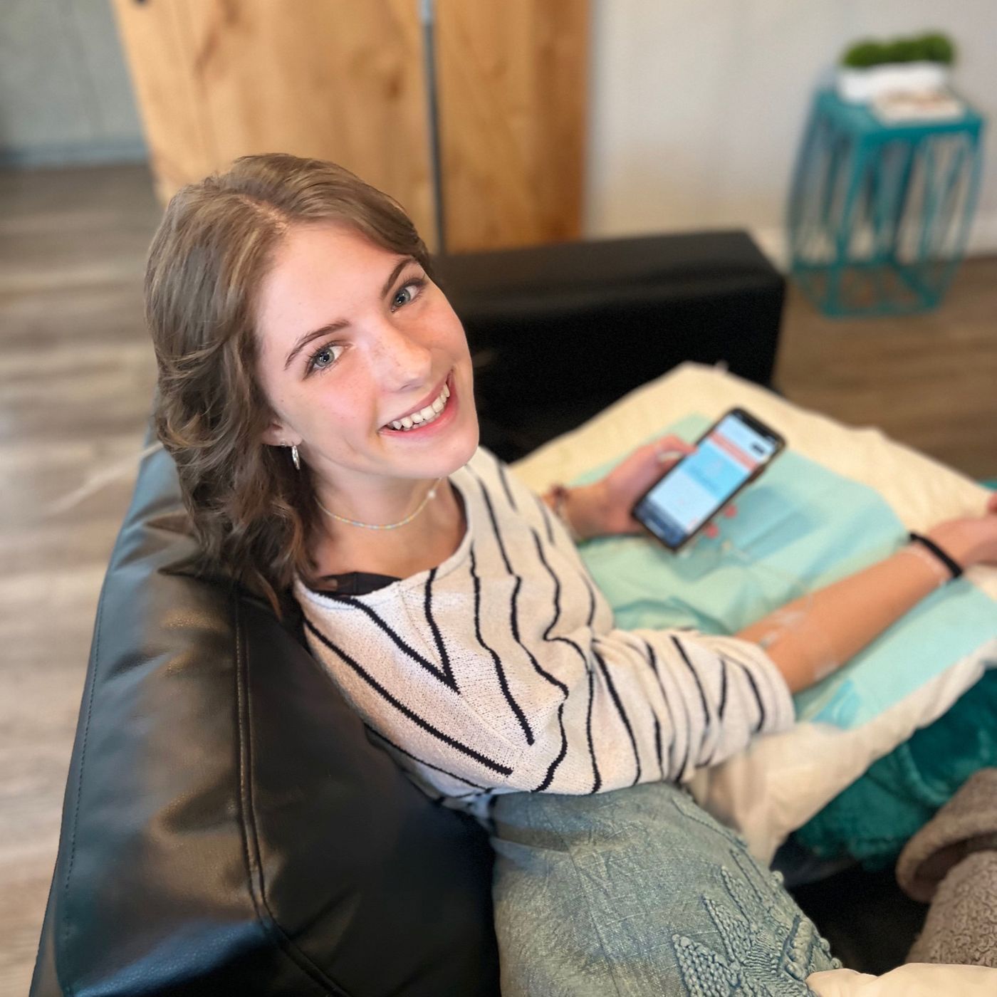 A woman is sitting on a couch holding a cell phone and smiling.