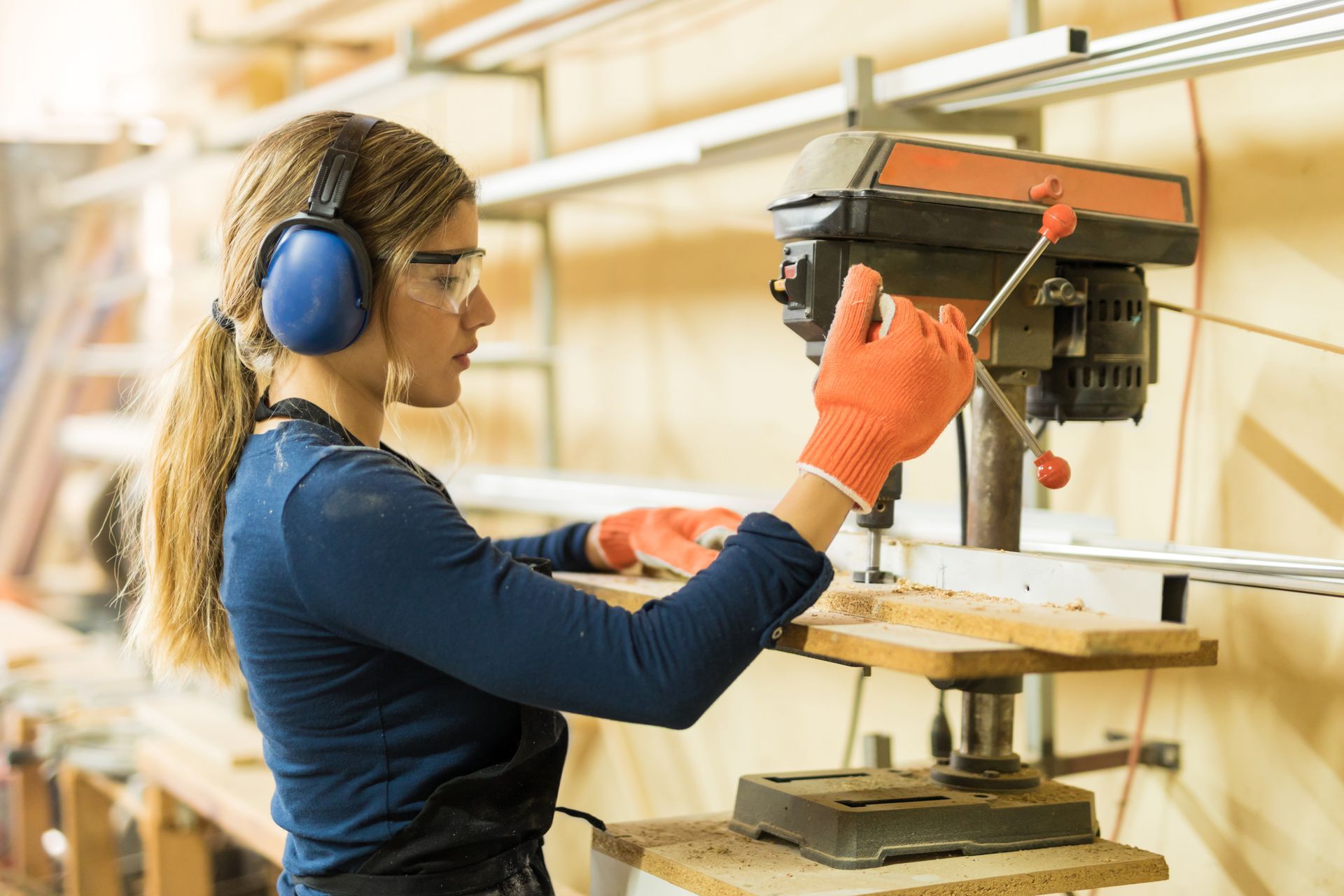 Person wearing blue earmuffs and orange gloves adjusts a drill press in a woodshop.