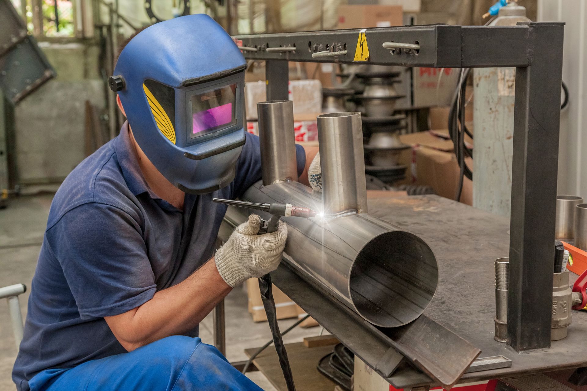 Worker in blue welding helmet welding a metal pipe in a workshop, with sparks flying.