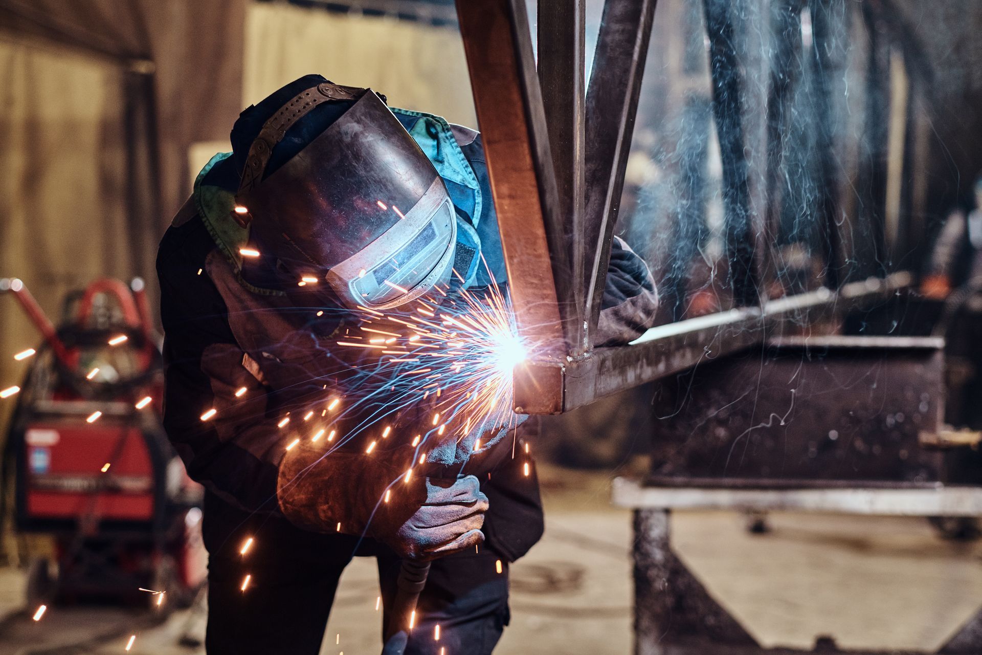 Welder in protective gear sparks metal in a workshop, with bright orange-blue arc light.