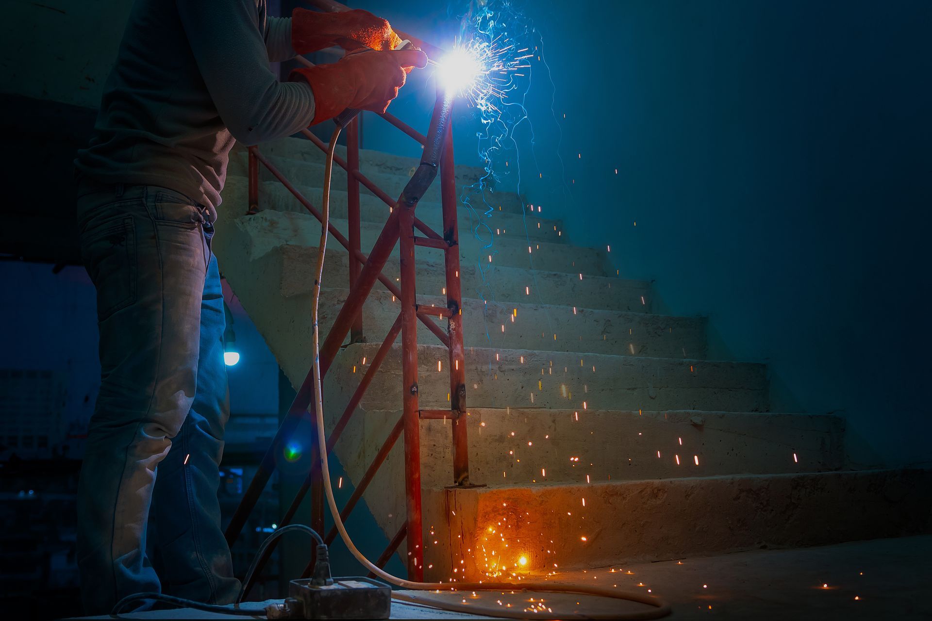 Welder in a dark stairwell, bright arc sparks flying as they weld a red metal railing