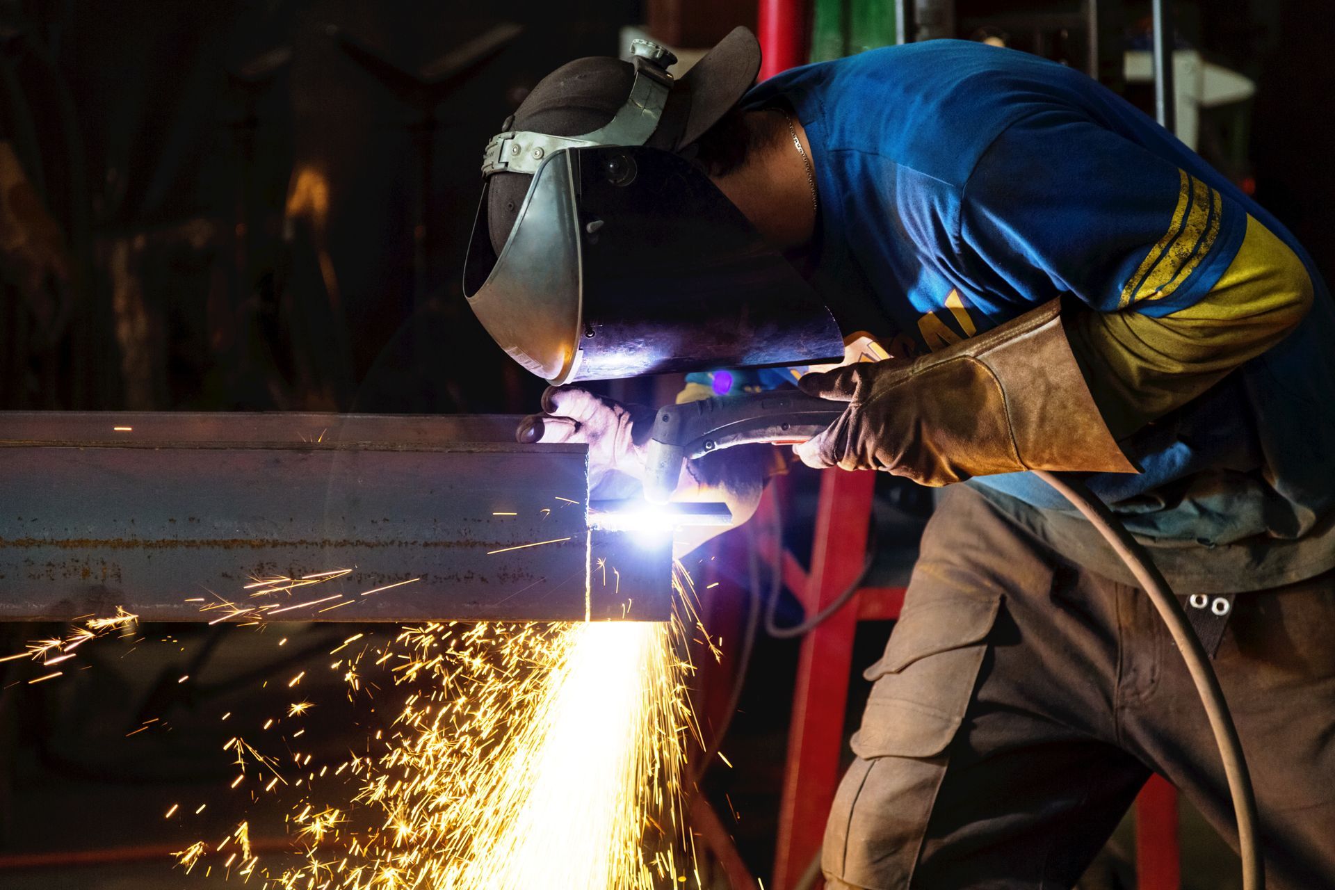 Welder in blue shirt and helmet sparks metal in a dark workshop