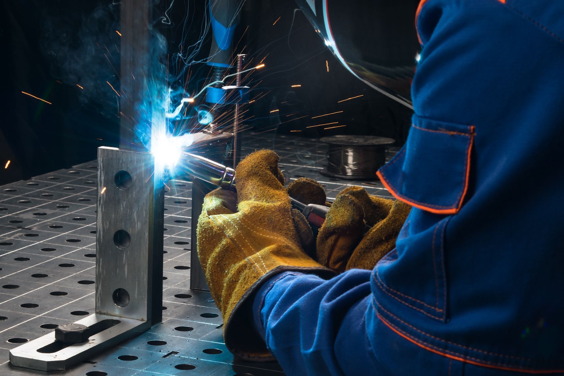Welder in blue protective gear welding a metal piece, with bright sparks in a workshop.