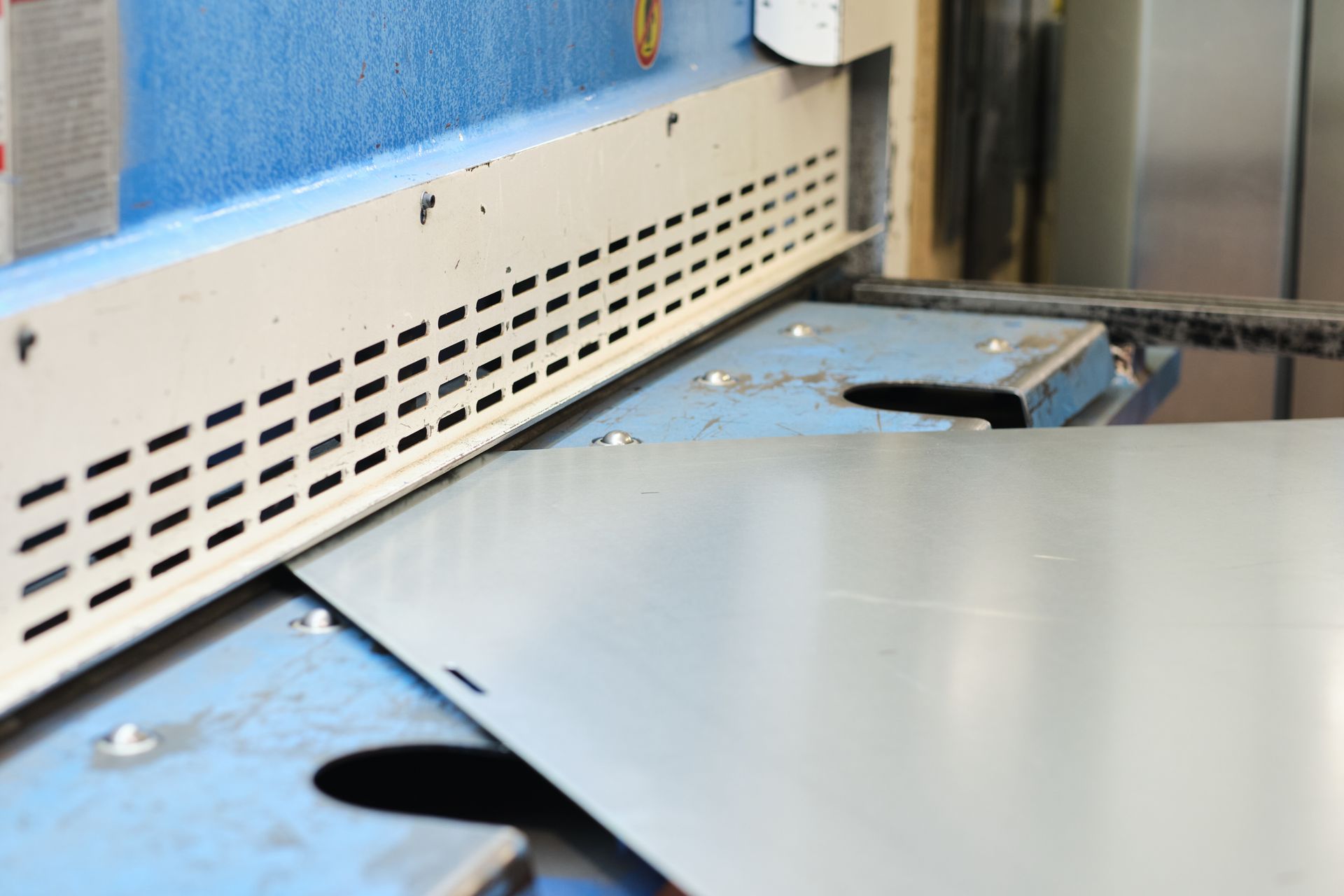 Silver metal sheet on a blue workbench beside a perforated vent panel in an industrial setting