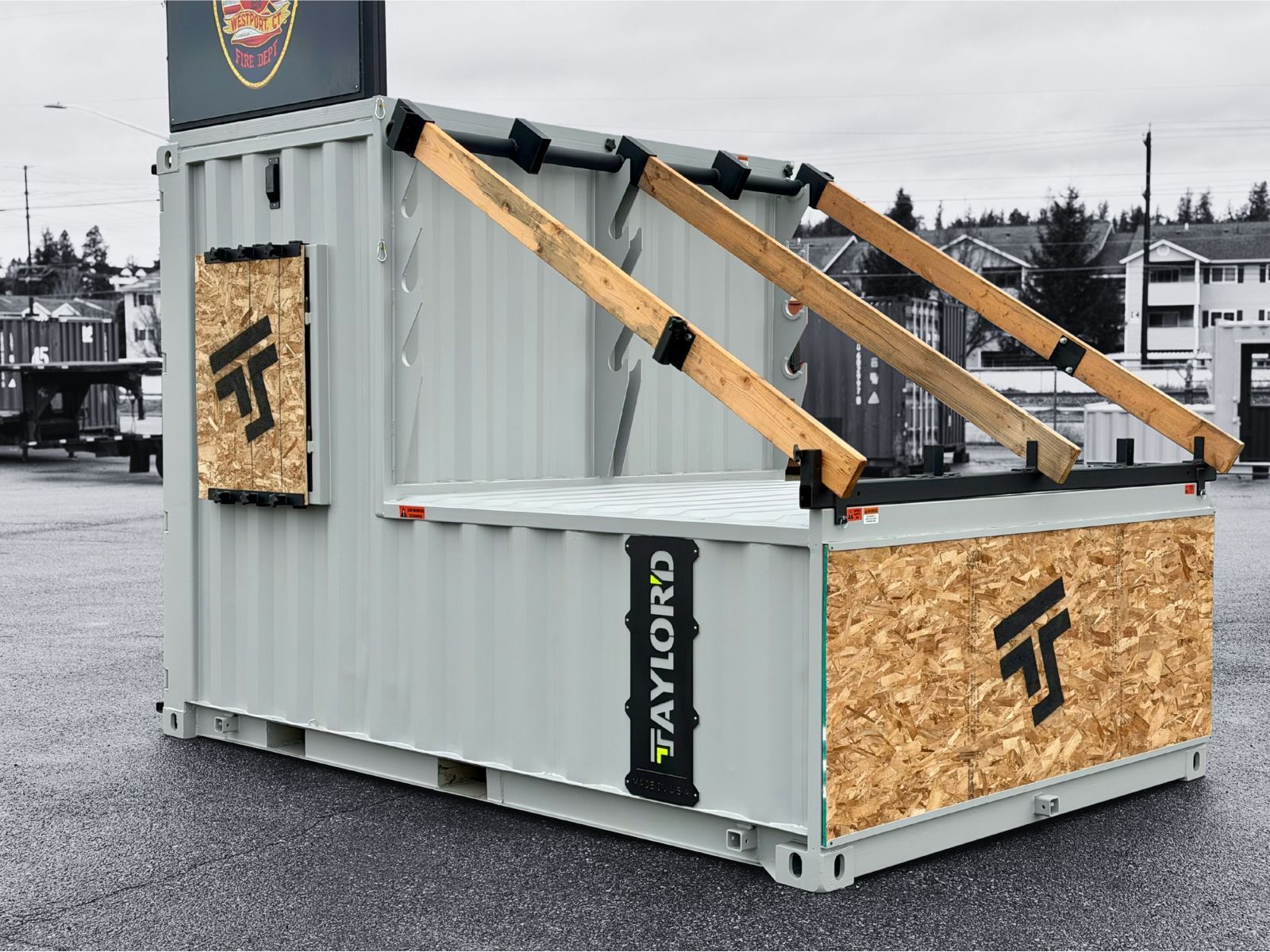 Modified shipping container with a ramp, and a rooftop platform. Gray and black. Outdoors.