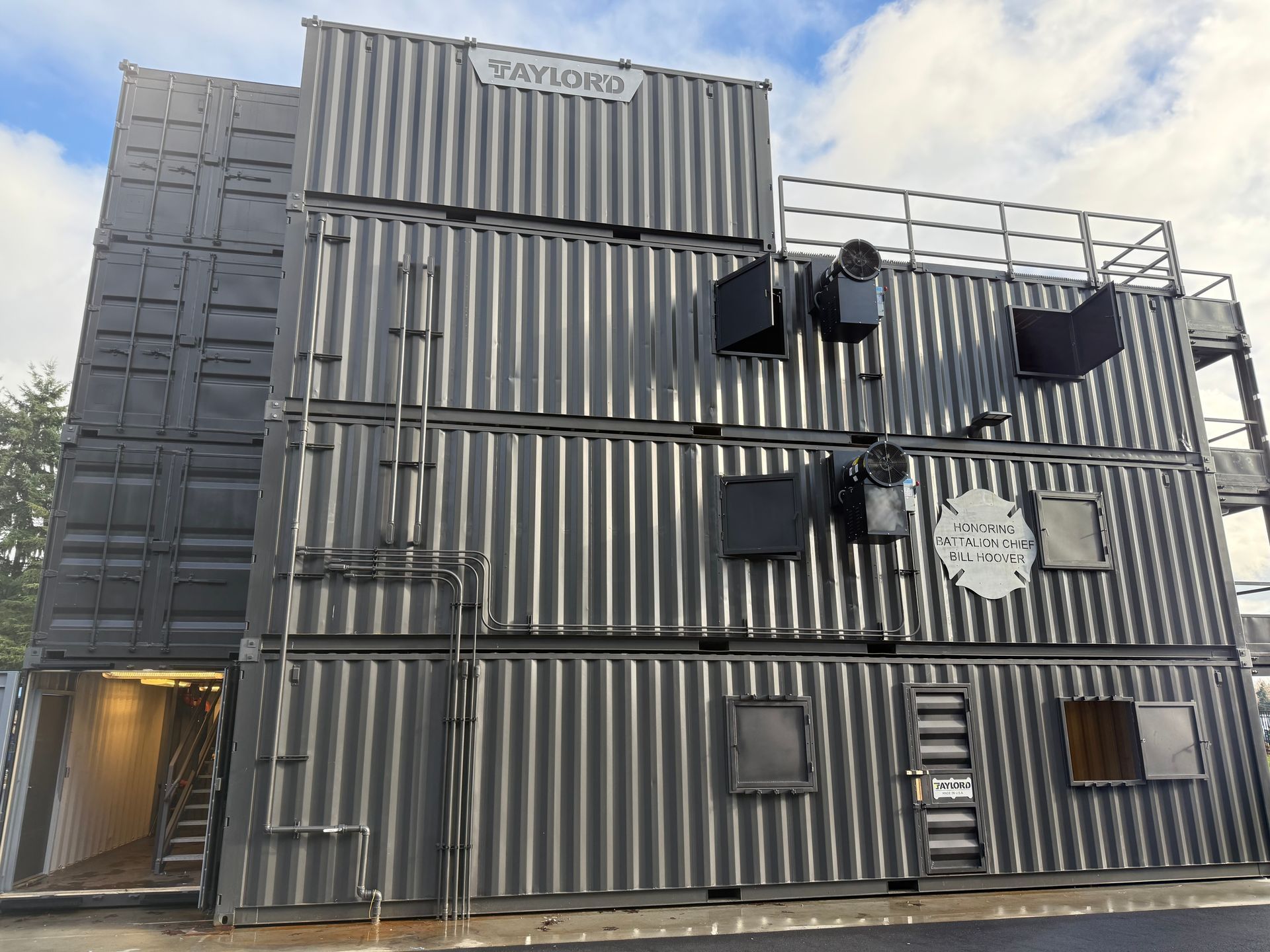 A multi-story structure made of stacked shipping containers, with doors, windows, and metal fixtures.