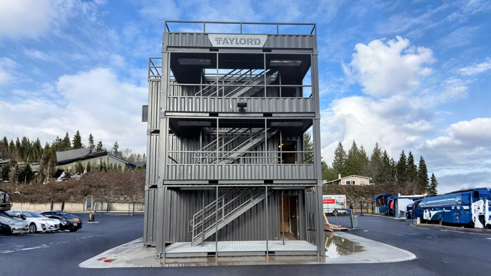 Three-story metal structure with exterior stairs, in a parking lot. Cloudy sky and trees in the background.
