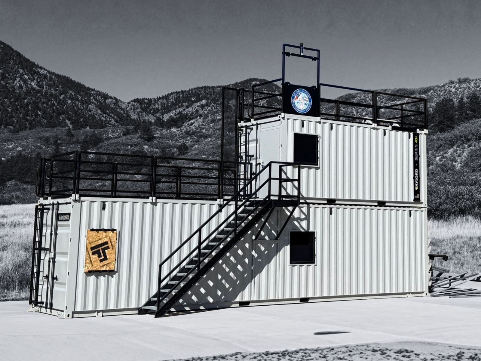 Two-story white shipping container structure with stairs, a rooftop platform, and logos, in a snowy field with mountains.