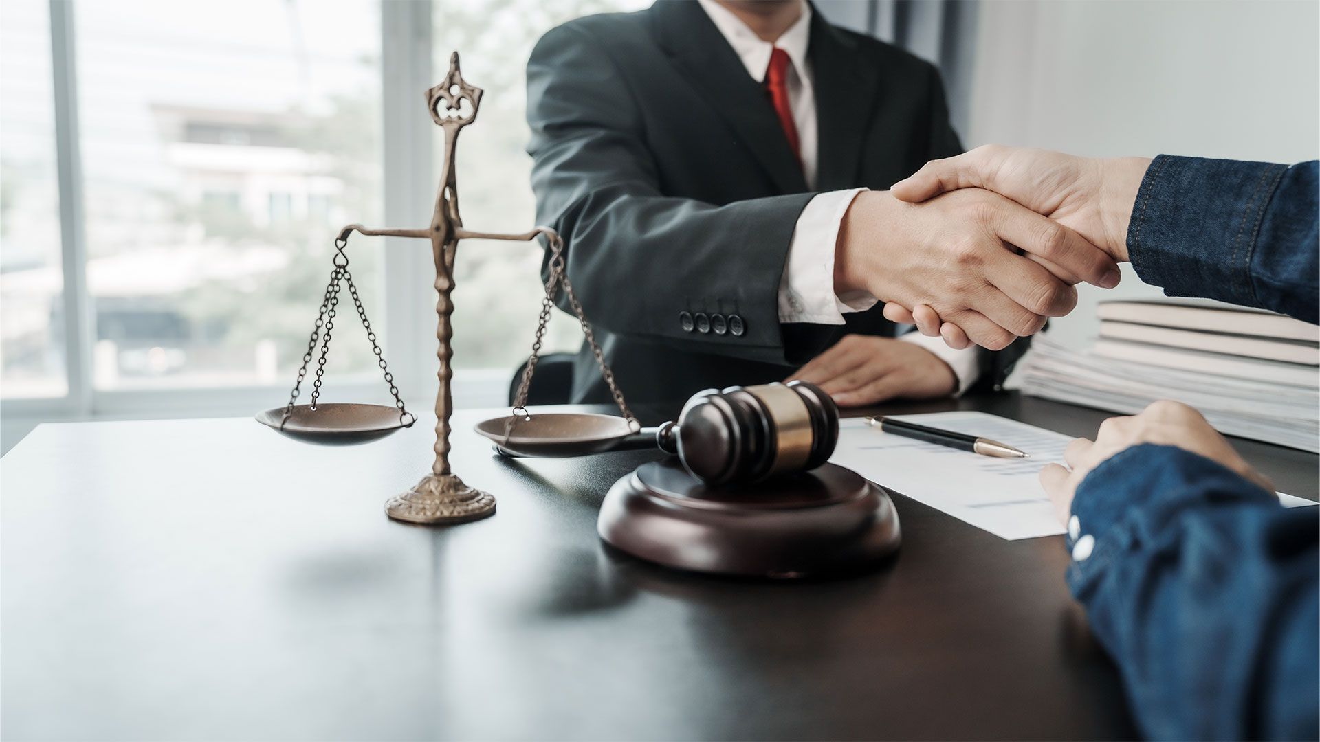Lawyer in a suit shaking hands with a client at a desk, with scales of justice and a gavel visible.