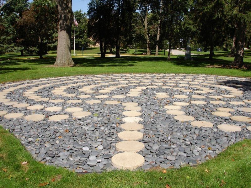 Stone labyrinth in a grassy park, with a circular path made of light-colored stones surrounded by dark gray pebbles.