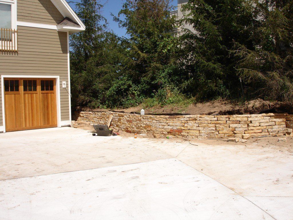 Concrete driveway next to a tan garage with a wooden door. A low stone wall sits in front of a hillside.