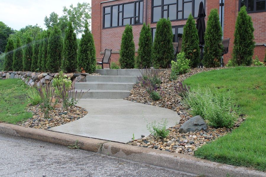 Concrete walkway and steps leading to a brick building, lined with greenery and rock landscaping.