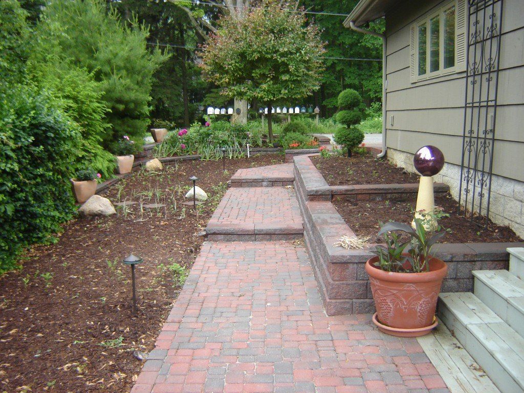 Brick pathway leads through a garden with raised beds and potted plants in front of a house.